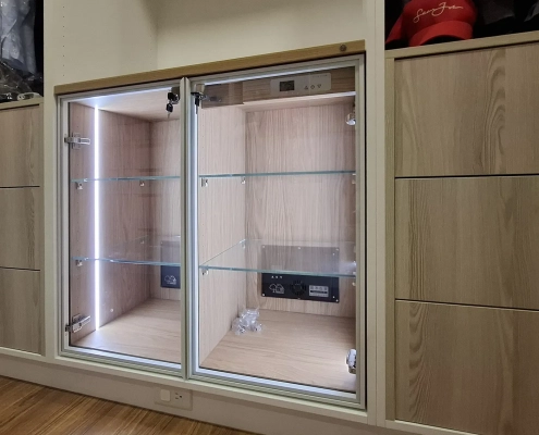 開放式更衣室中央衣櫃下層訂製嵌入除濕防潮精品展櫃完工照，兩側為既有木質抽屜，中央配置細鋁框對開玻璃門、垂直氛圍燈條、通透玻璃層板與獨立除濕模組。
