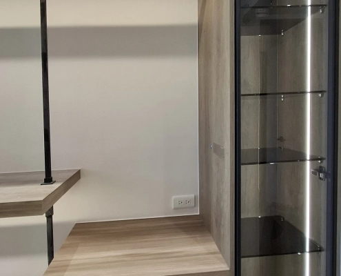 更衣室中景視角，展示天花板樑下黑鐵吊衣架與下層木紋抽屜收納平台，轉角銜接極窄黑鋁框灰玻除濕防潮高櫃，右側透出灰玻層板與直立式 LED 光帶，透過自主研發除濕模組在開放空間中打造 45%～55% RH 獨立防潮微氣候。
